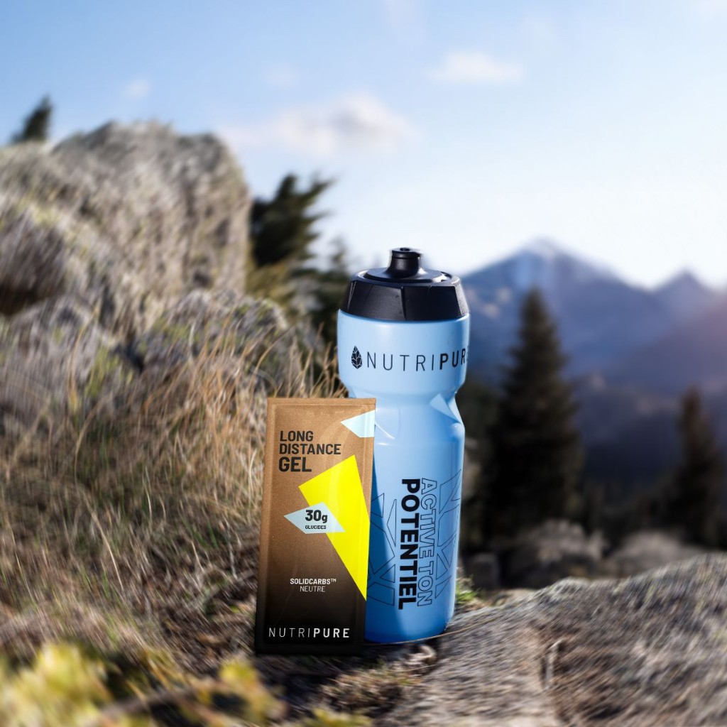 Gel longue distance et gourde Nutripure sur un rocher, arrière-plan montagnard flou (visuel produit, nutrition sportive).